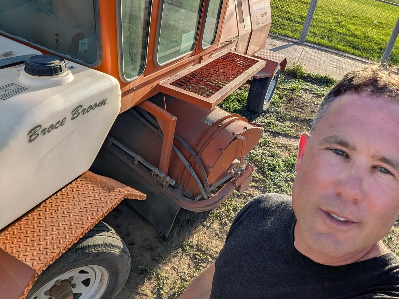 Rhett Crites beside the water tank and operator platform of an orange Broce Broom road sweeper, on grass in bright sun.
