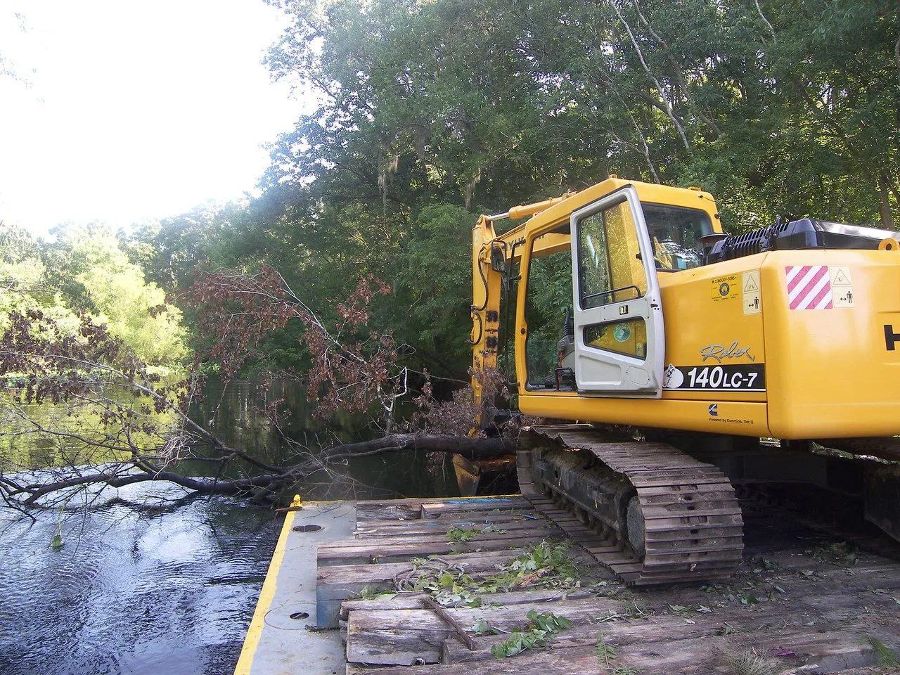 A yellow Hyundai Robex 140LC-7 excavator, its cab door open, parked on the timber-matted deck of a barge on the Ocklawaha River beside a fallen tree lying in the water.