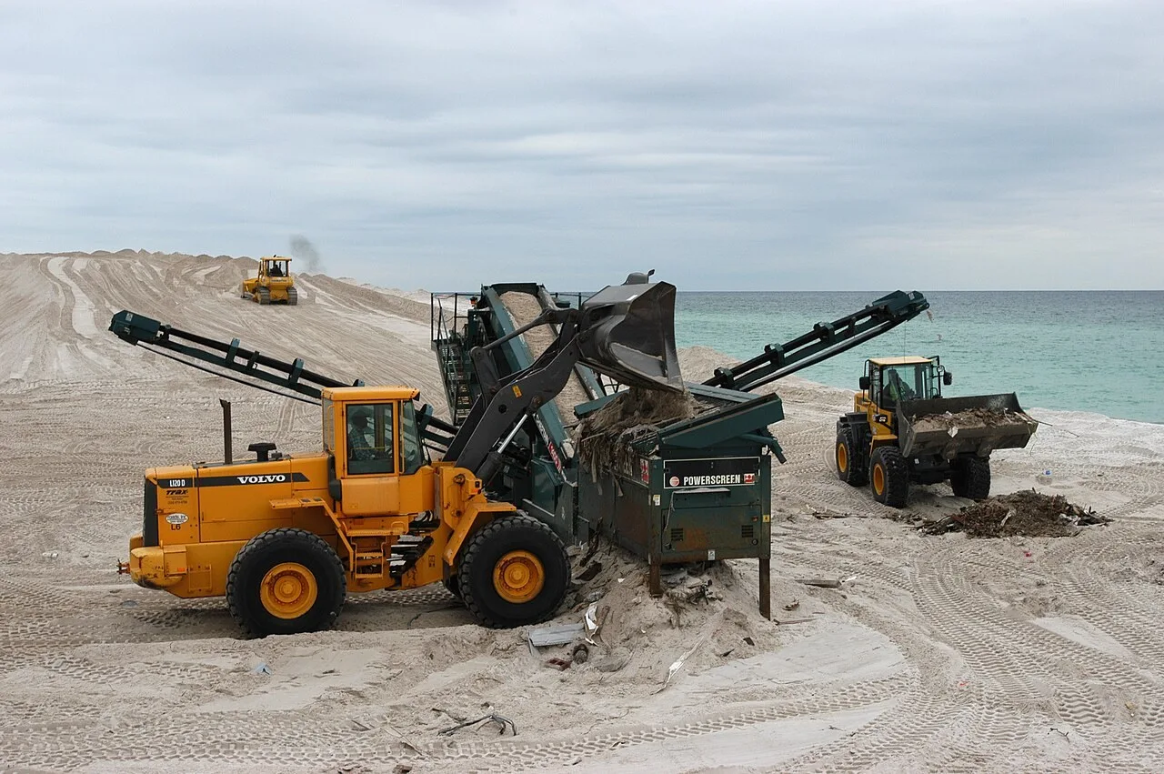 A Volvo L120D wheel loader tips sand and debris into a Powerscreen screening plant on Pensacola Beach, Florida, with a second wheel loader beside the water and a bulldozer on the dune behind.