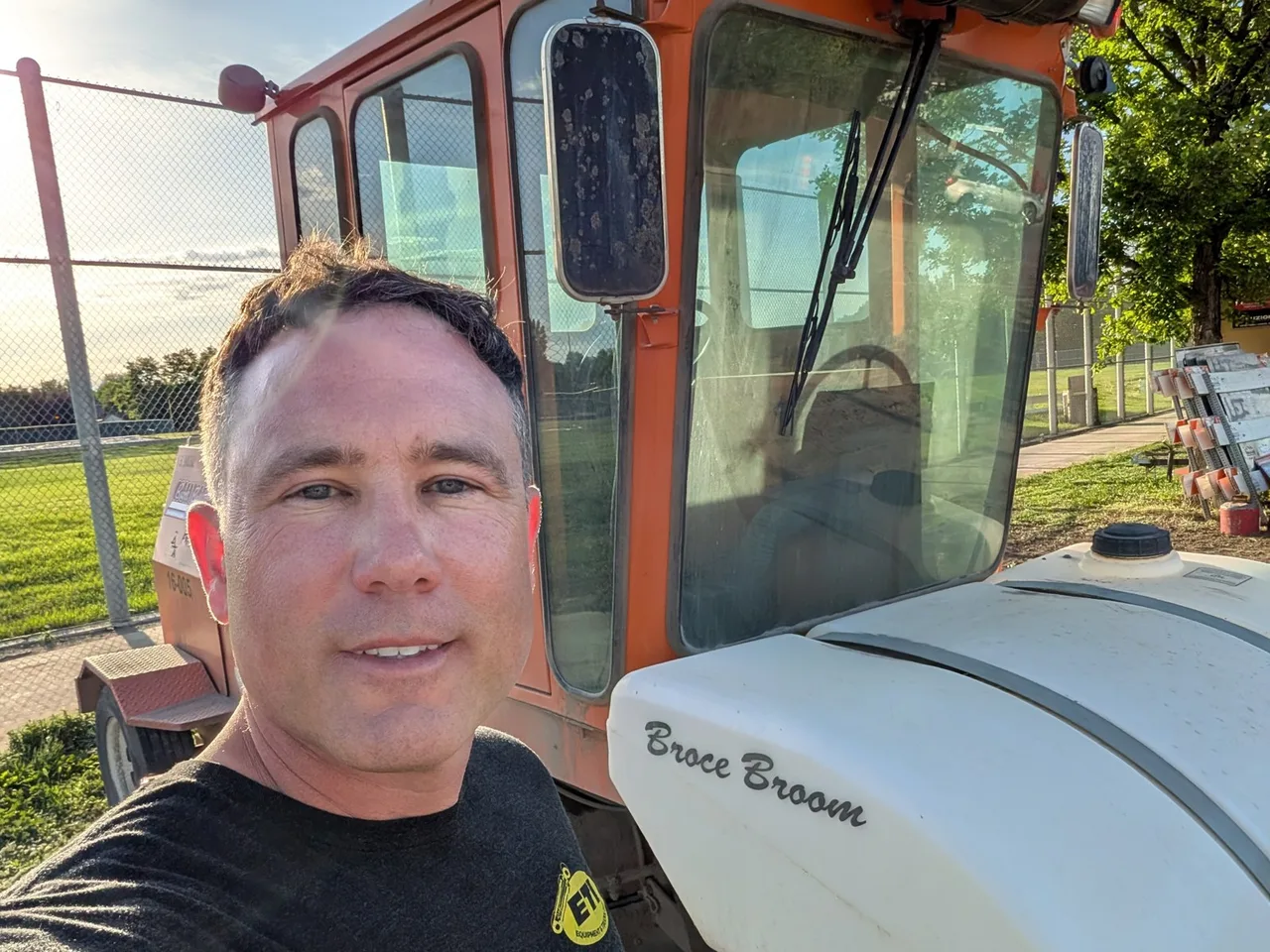 Rhett Crites in front of an orange Broce Broom road sweeper on a sunny day, with green grass and trees behind.