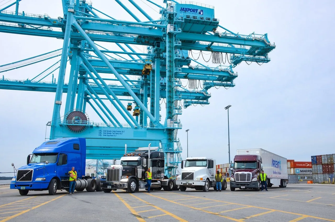 Four semi trucks lined up in front of a blue JAXPORT container crane in Jacksonville, Florida, with stacked shipping containers behind them.
