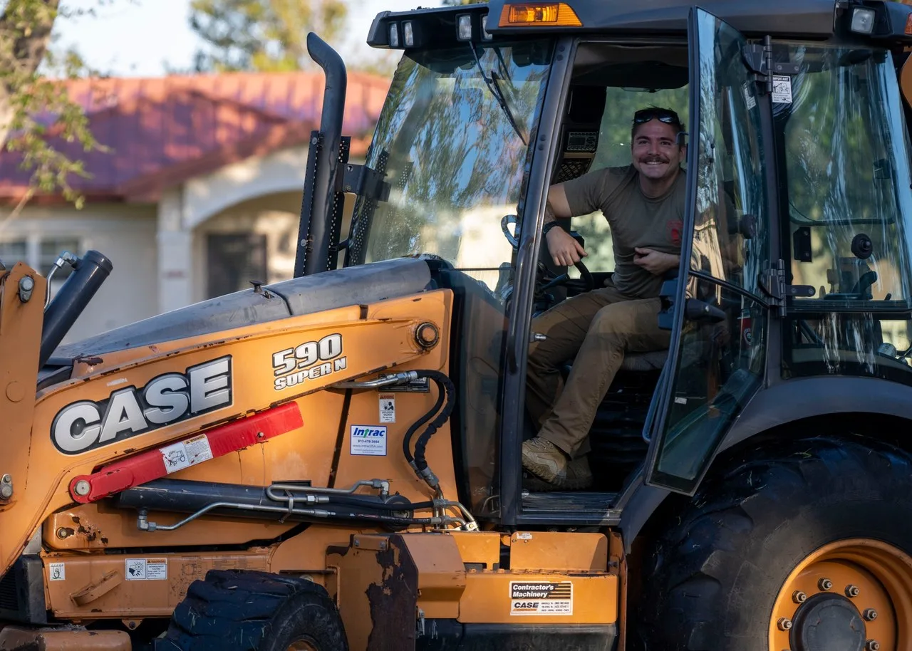 An orange Case 590 Super N backhoe loader at MacDill Air Force Base, Florida, with its operator in the open cab door and a red-roofed house behind.