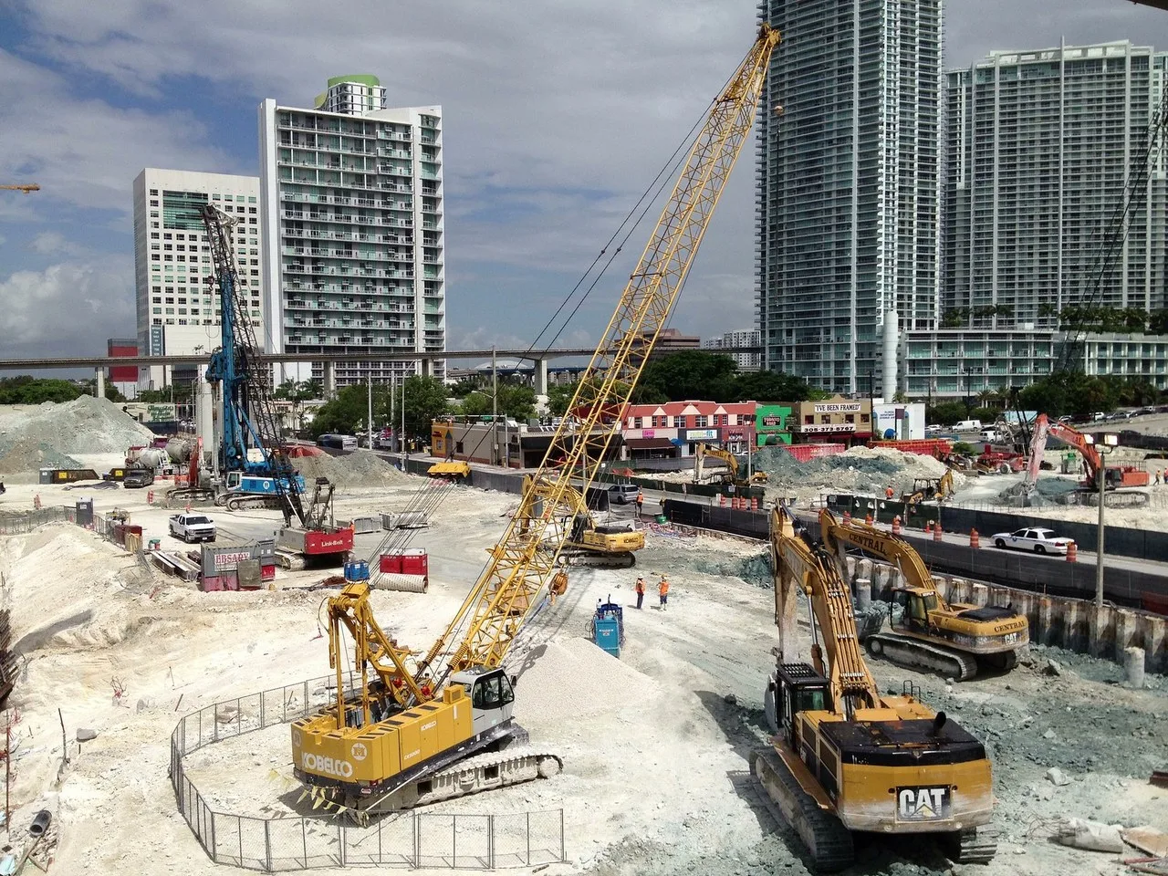 A yellow Kobelco crawler crane, a blue piling rig and two Cat excavators work a rock pit at the Brickell CityCentre construction site, with high-rise towers behind them.