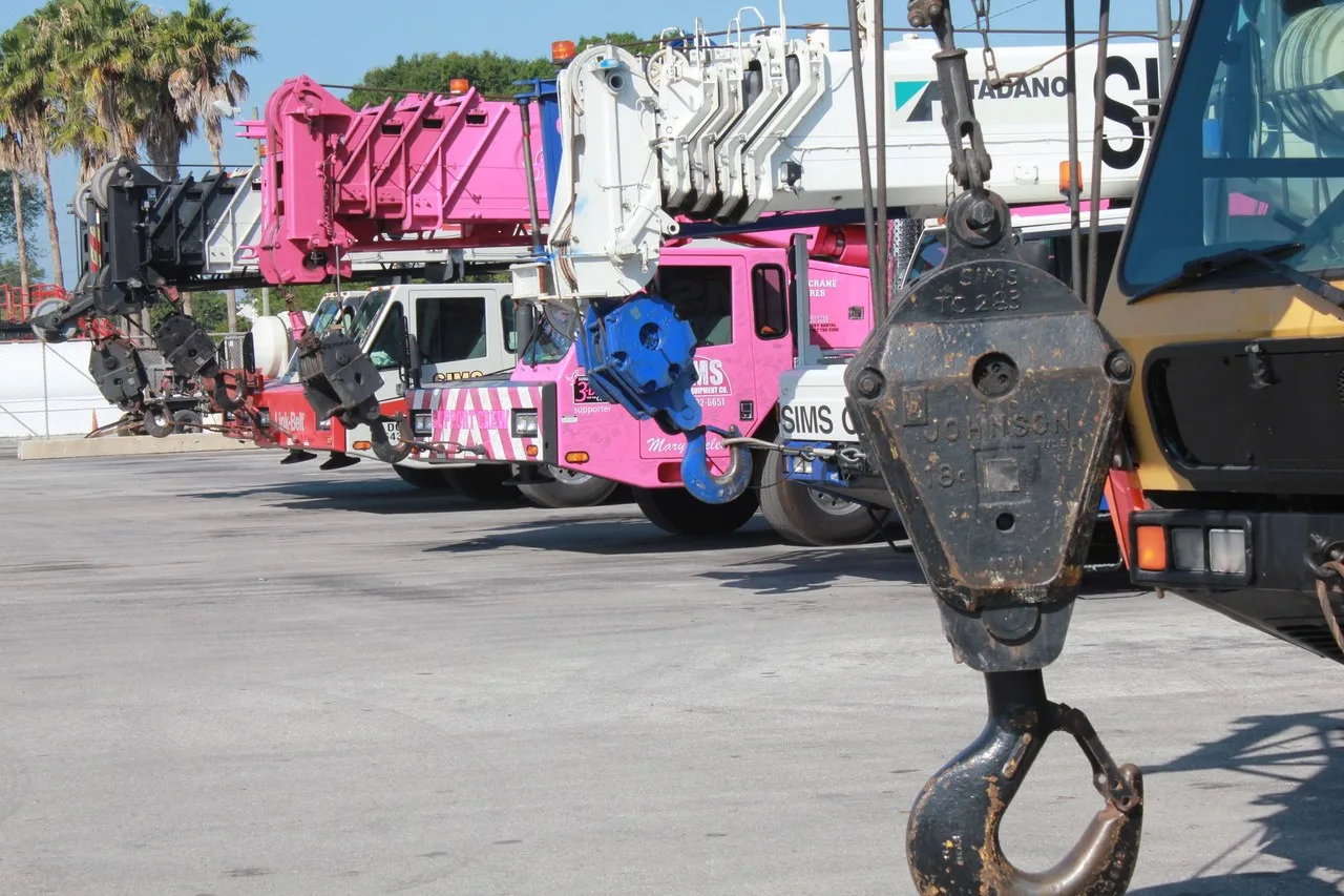 A row of telescopic truck cranes, one pink and one white with a Tadano boom, parked on a paved lot in Tampa, Florida, with a hook block hanging in the foreground and palms behind.