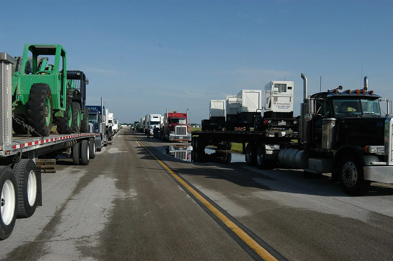 Semi trucks with flatbed trailers, one carrying generator sets and another a green rough-terrain machine, lined up at Lakeland Linder Regional Airport in Lakeland, Florida.
