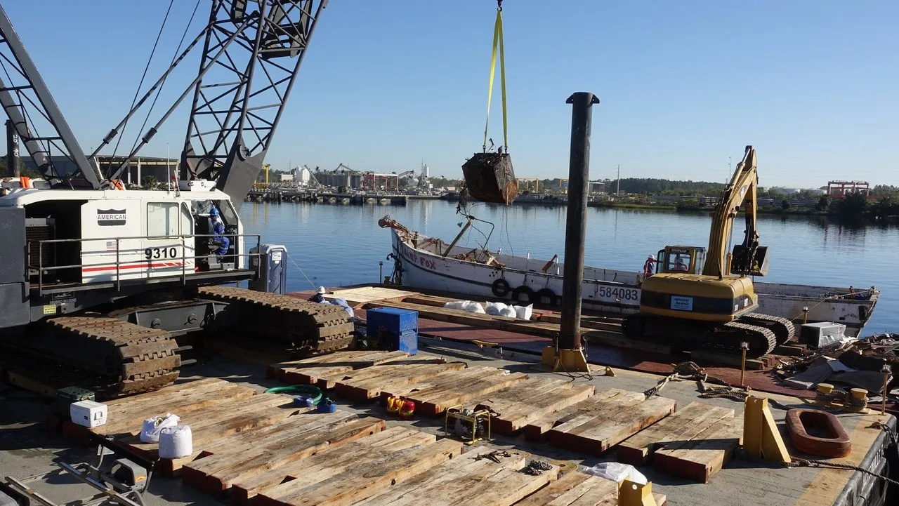 An American 9310 crawler crane and a tracked excavator working from a timber-matted barge on the Trout River in Jacksonville, Florida.