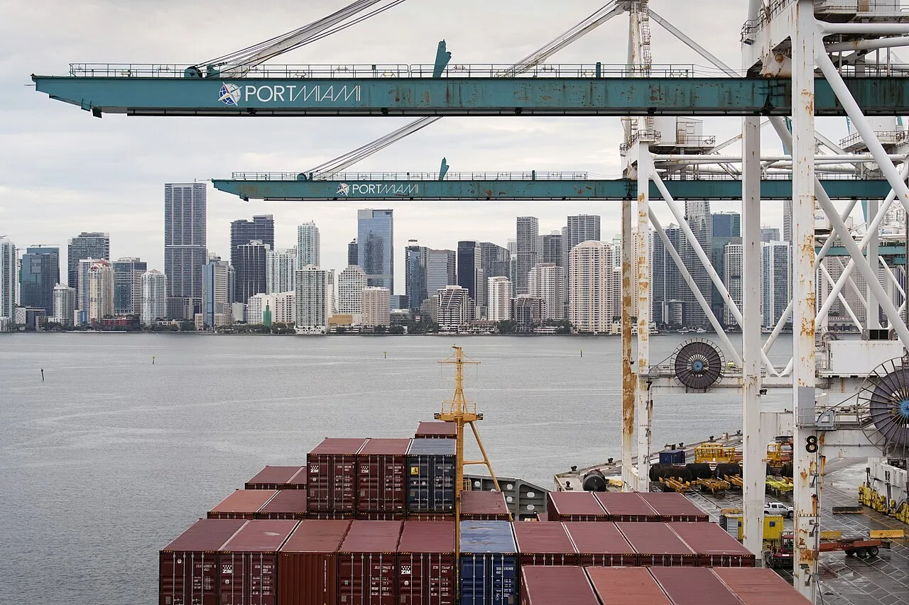 Teal ship-to-shore crane booms lettered PORTMIAMI reach over the stacked containers of a cargo ship docked at PortMiami in Miami, Florida, with the Miami skyline across the bay.