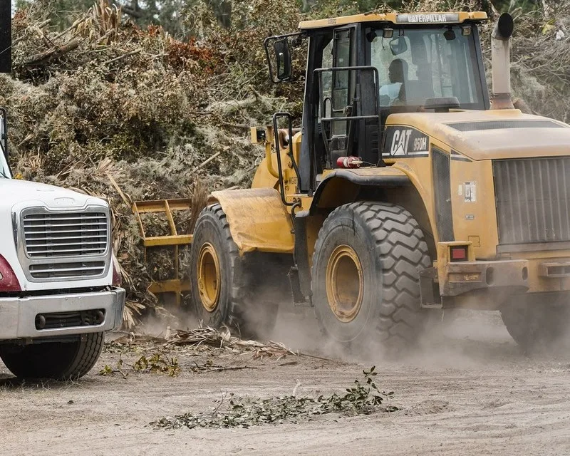 A yellow Cat 950H wheel loader with an attachment on its front works beside a pile of storm debris at a debris site in Sanford, Florida, with the front of a white truck at the left edge.