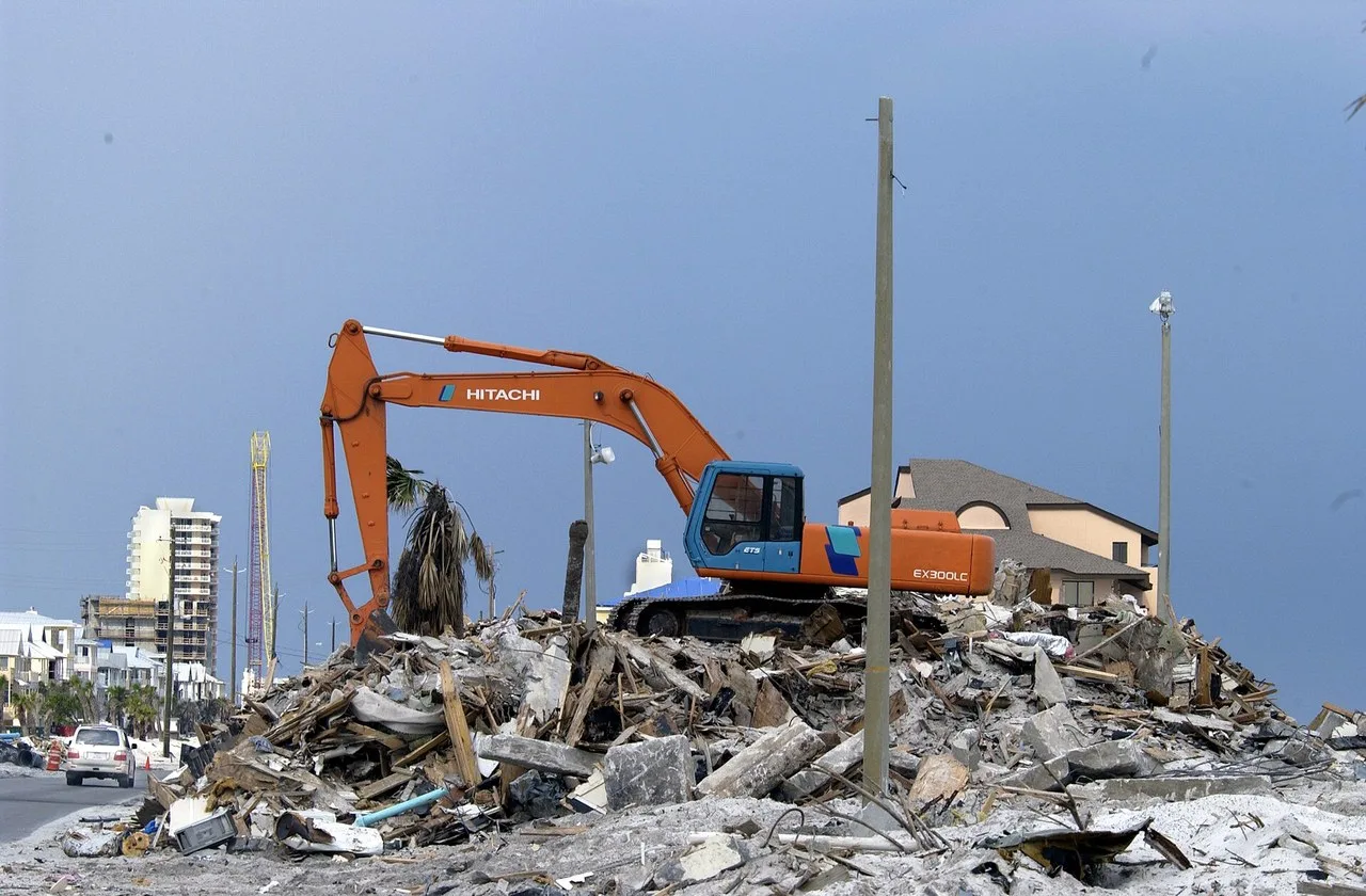 An orange Hitachi EX300LC excavator sits on a pile of construction debris at Pensacola Beach, Florida, with palms and beachfront buildings behind it.