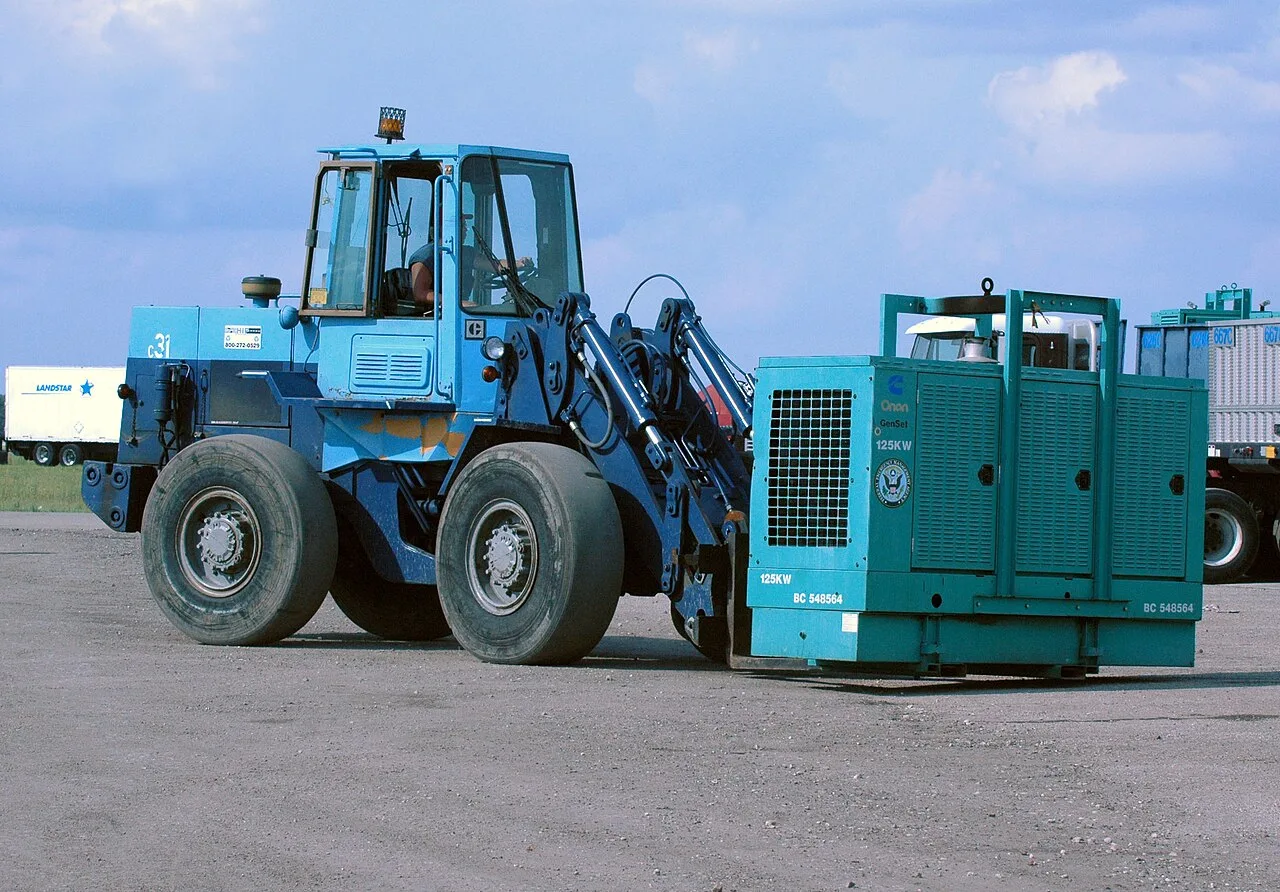 A blue wheel loader fitted with forks carries a Cummins Onan 125 kW generator set across a staging area in Lakeland, Florida.
