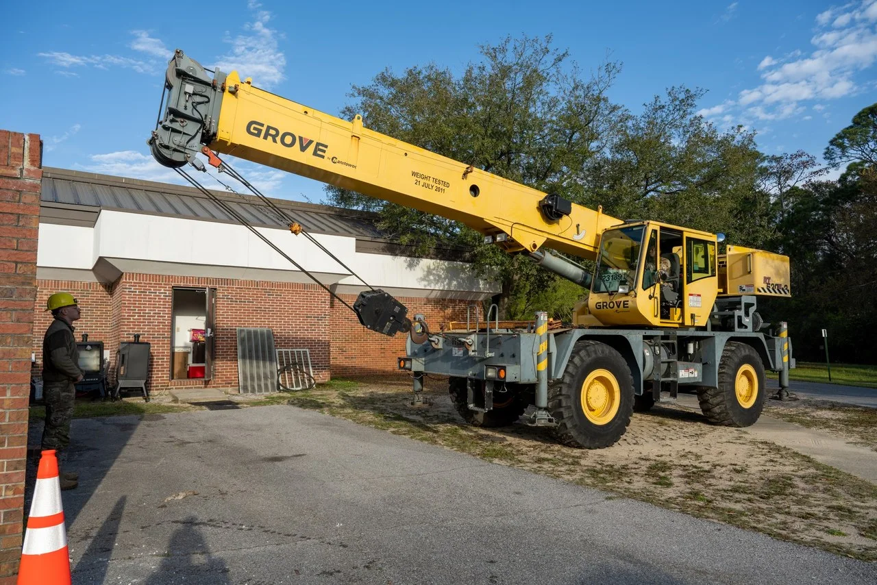 A yellow Grove rough-terrain crane with its boom extended beside a brick building at Hurlburt Field, Florida, while a worker in a hard hat looks on.