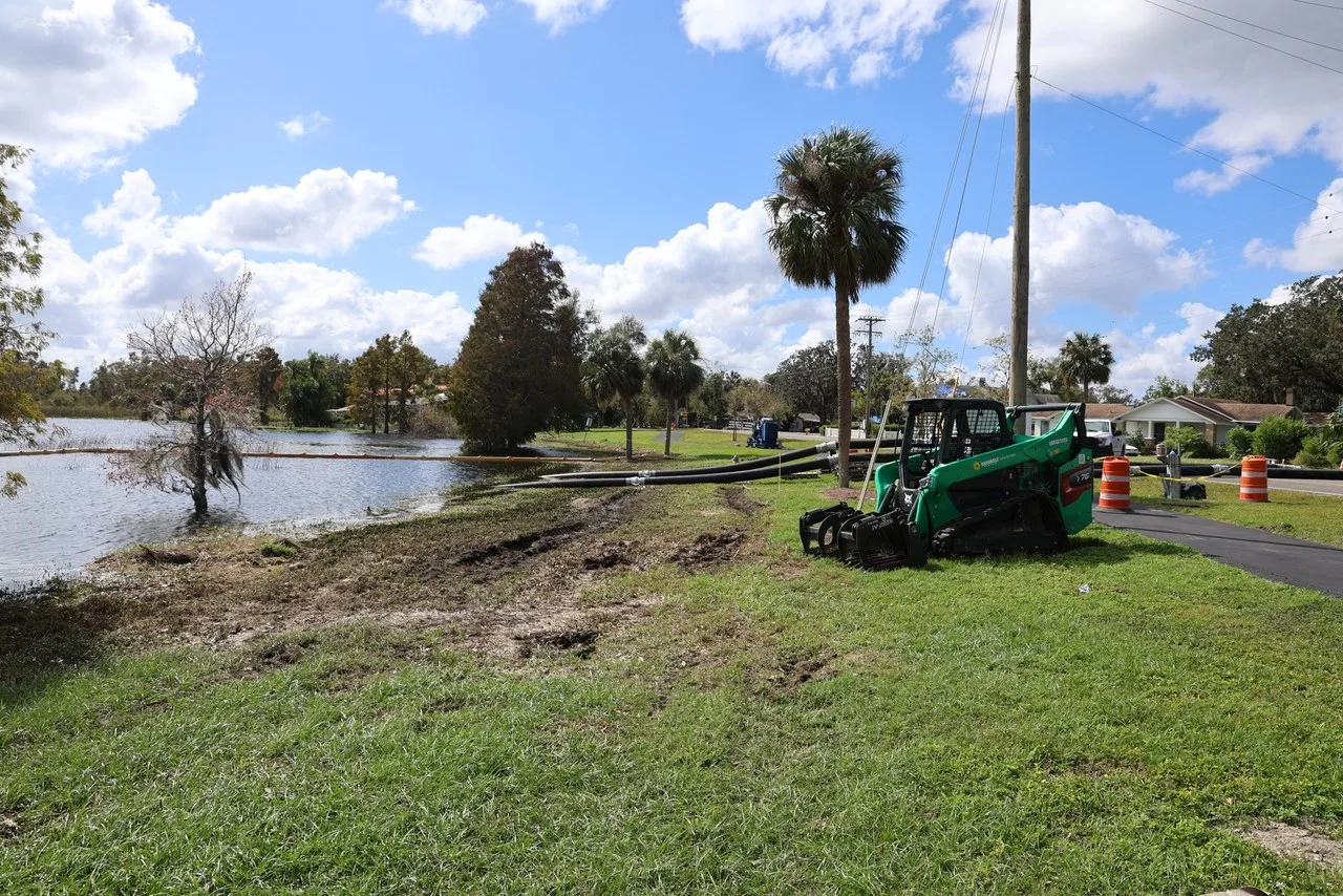 A green compact track loader with a grapple attachment sits beside pump hoses on the shore of Lake Bonny in Lakeland, Florida.