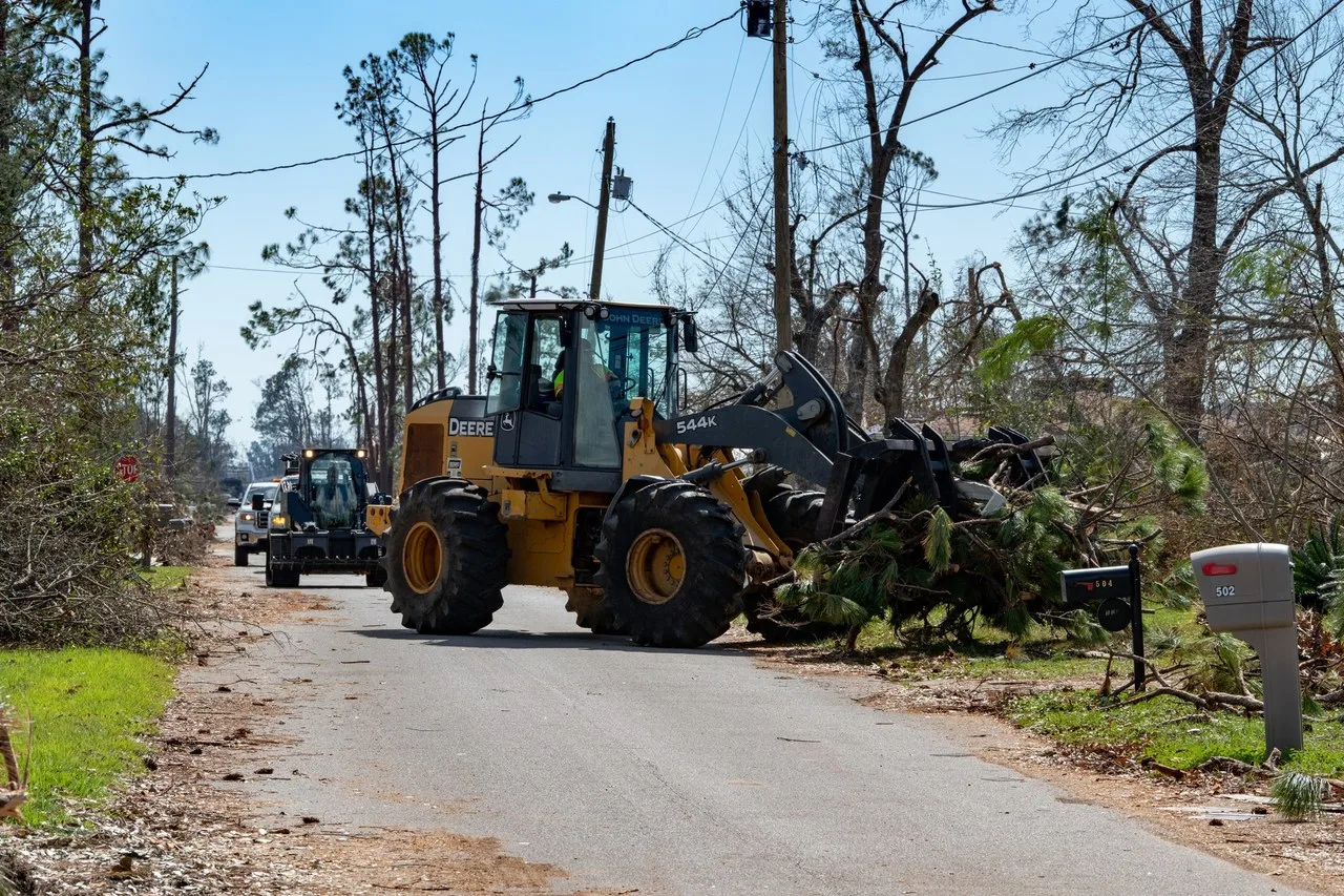 A John Deere 544K wheel loader with a grapple pushes fallen pine debris along a street in Lynn Haven, Florida, with a second machine and a pickup truck behind it.