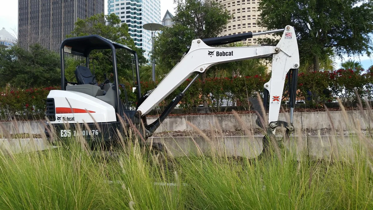A white Bobcat E35 compact excavator with its boom stretched over a low concrete wall, seen through tall grass, with downtown Tampa, Florida, office towers behind.