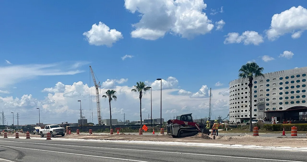 A red compact track loader and two workers in safety vests on road work along Universal Boulevard in Orlando, Florida, with palms and a lattice-boom crane behind them.