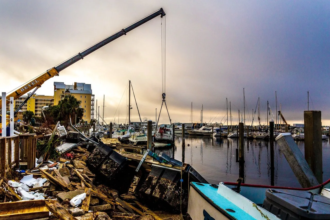 A yellow telescopic crane boom lifts a boat from the water at St. Andrews Marina in Panama City, Florida, with storm debris and wrecked docks in the foreground and moored boats behind.