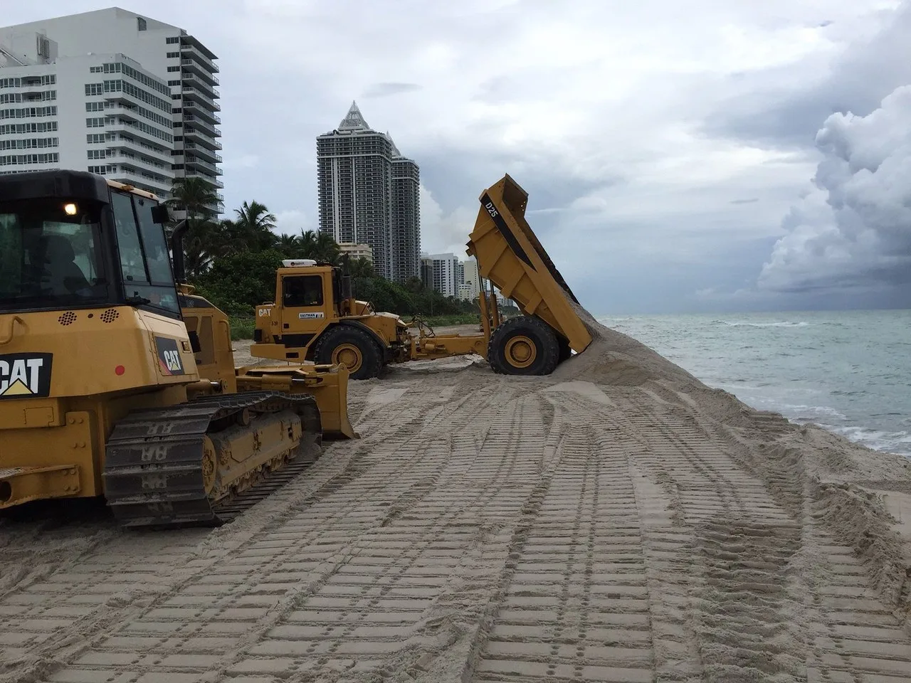 A Cat bulldozer in the foreground and a yellow articulated dump truck tipping sand at the water’s edge during beach renourishment at Miami Beach, with oceanfront towers and palms behind.