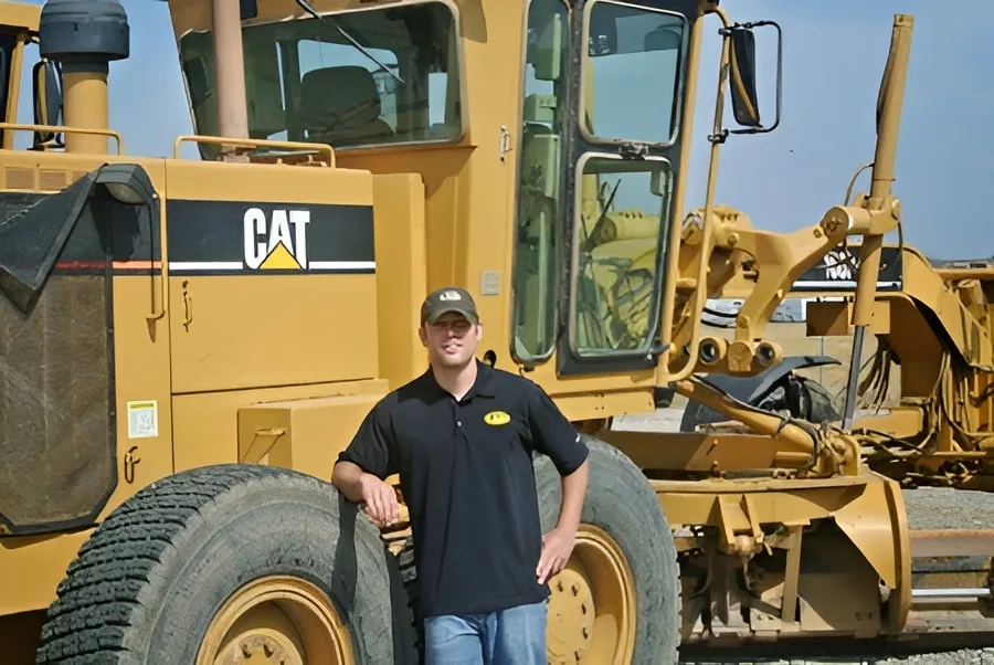 Rhett Crites early in his career, leaning on the front tire of a Cat motor grader in an equipment yard.