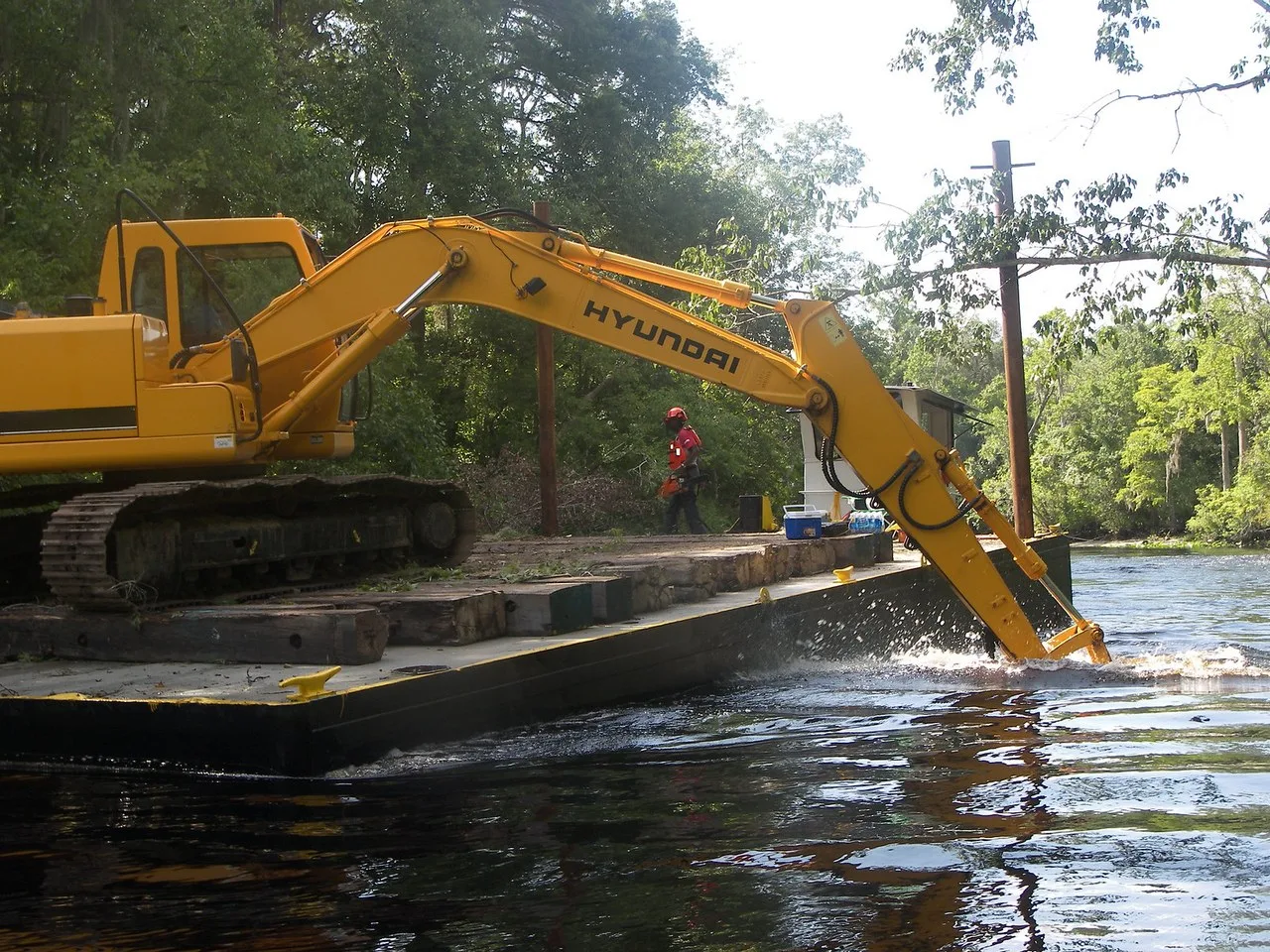 A yellow Hyundai crawler excavator on a deck barge reaches its boom into the Ocklawaha River, with a worker in a red hard hat on the barge and trees along the bank.